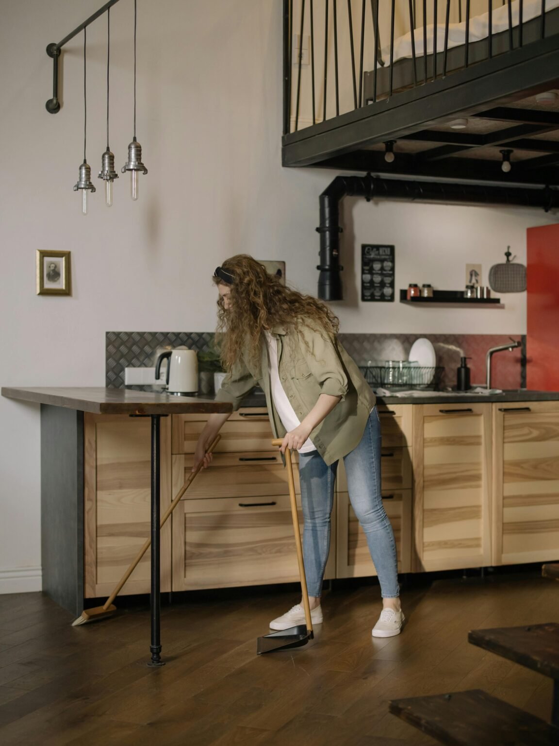 Young woman cleaning a modern loft kitchen area with a broom, demonstrating home maintenance.